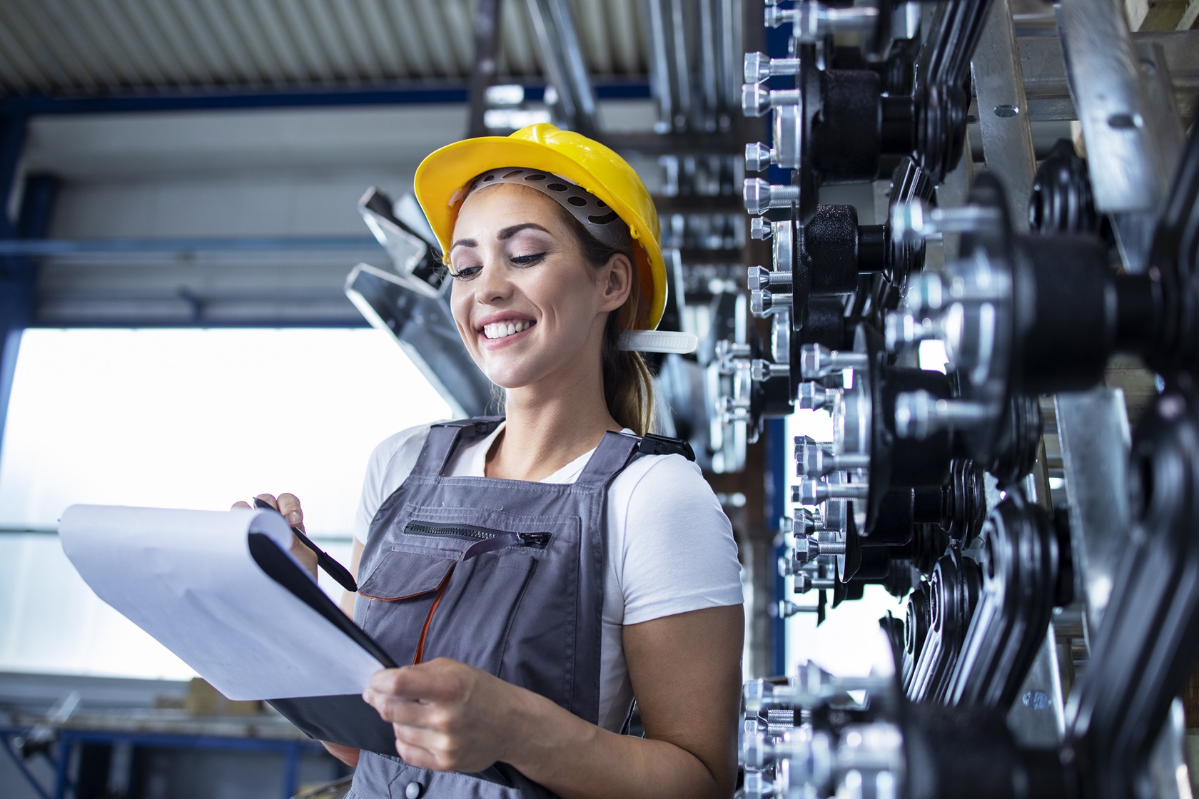 Portrait of female industrial employee in working uniform and hardhat writing production results in factory.